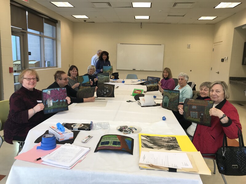 A painting workshop facilitated by Patricia Dubroof, shows a group of people seated around a table, holding up their completed paintings