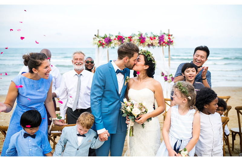 Los recién casados ​​se besan después de su hermosa boda en la playa, mientras amigos y familiares lanzan pétalos de flores y vitorean