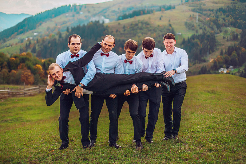 Los padrinos de boda se paran en fila en una colina al aire libre, sosteniendo al novio en una pose tonta