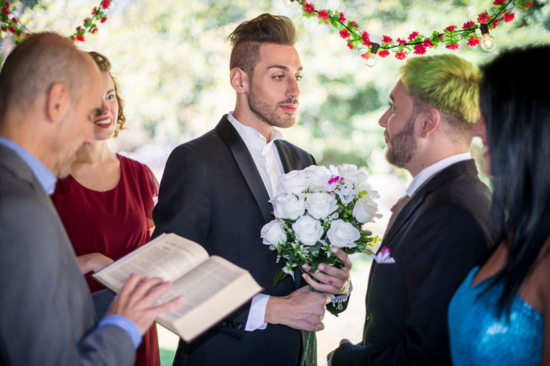 Officiant leads the wedding ceremony for two grooms on a sunny day as friends and family members watch.