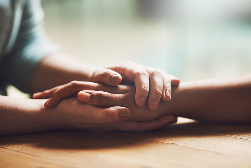 Close up photo shows two people holding hands in a loving way, resting hands on tabletop