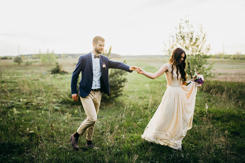 Newlyweds hold hands in a field on their wedding day, the scenery feels ethereal, romantic, and dreamy