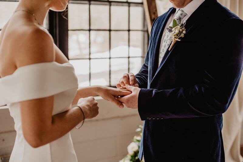 Bride and groom hold hands in front of a window, dressed in their formal wedding attire