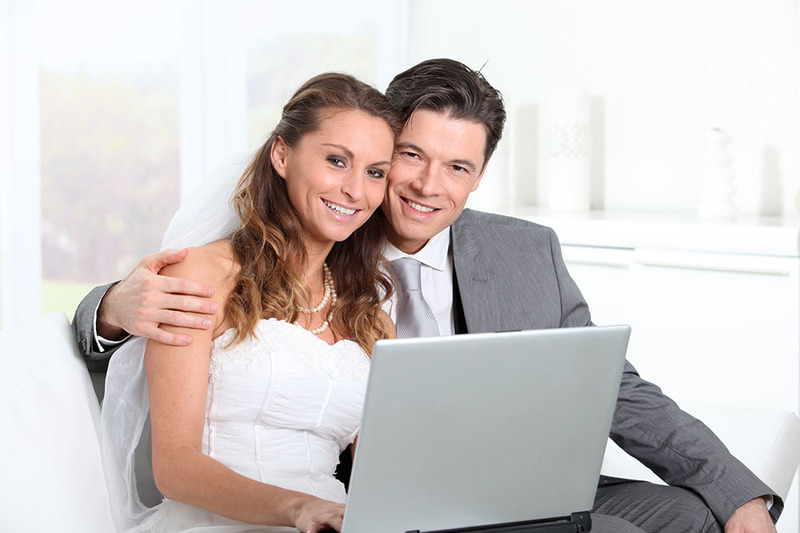Bride and groom embrace on the wedding day, holding a laptop for their remote online wedding ceremony