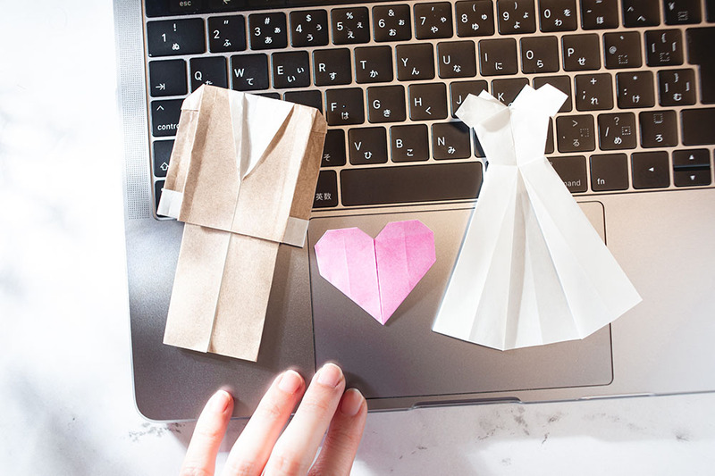 Paper cut outs of a bride's wedding dress and groom's suit are placed on a laptop keyboard, symbolizing online wedding ceremonies