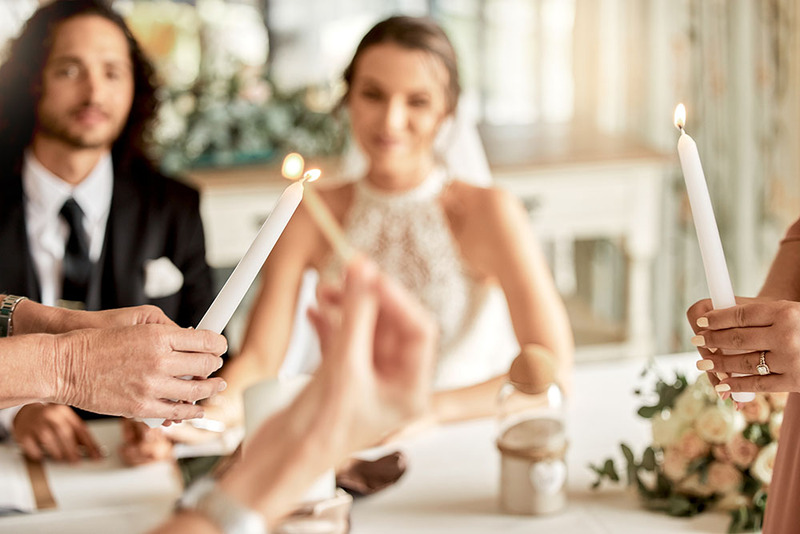 In the background, a groom and bride sit at a table surrounded by flowers and beautiful wedding decor. In the foreground, two women hold candles to perform the unity candle ceremony, mother's helping with the ritual