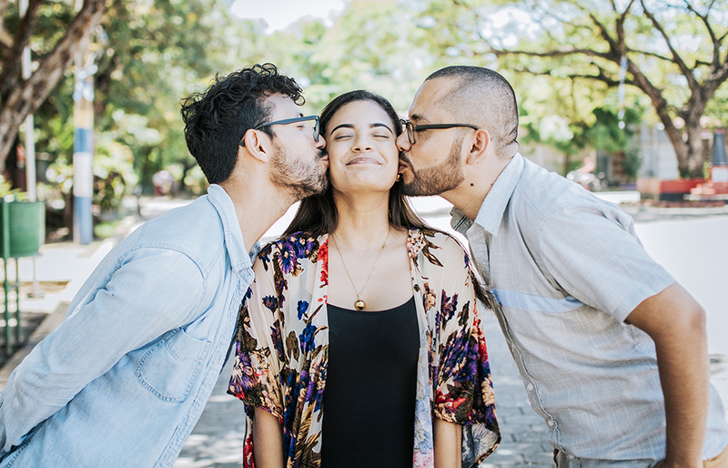A group of three poly partners, two partners stand on either side of the third and kiss her cheeks. She smiles happily with her eyes closed. The photo is sweet, wholesome looking, cute. They are outdoors in the sun with trees behind them.