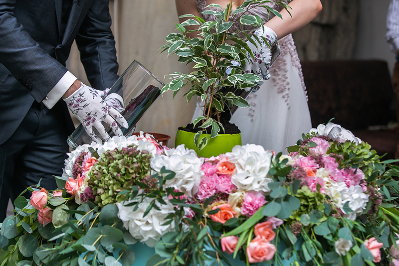 Close up of a groom and bride planting a tree in a small container during a wedding ceremony. They are wearing formal wedding suit and dress and cute gardening gloves with flowers