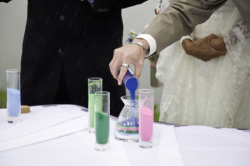 Close up shows a groom pouring colored sand into a clear vase during a unity sand ceremony, other partners watch in the distance