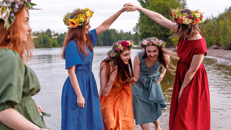 Women with flower head wreaths are wearing bright colors and laughing and dancing on the wedding day. Behind them is a lake, and in the distance a forest