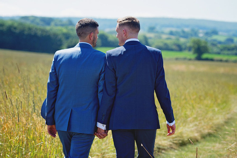 Two grooms walk holding hands in a field on the wedding day in Virginia, in the distance are mountains and trees