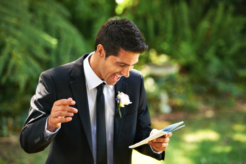 A man in a suit reads from a wedding script while officiating an outdoor garden wedding. He gestures and smiles