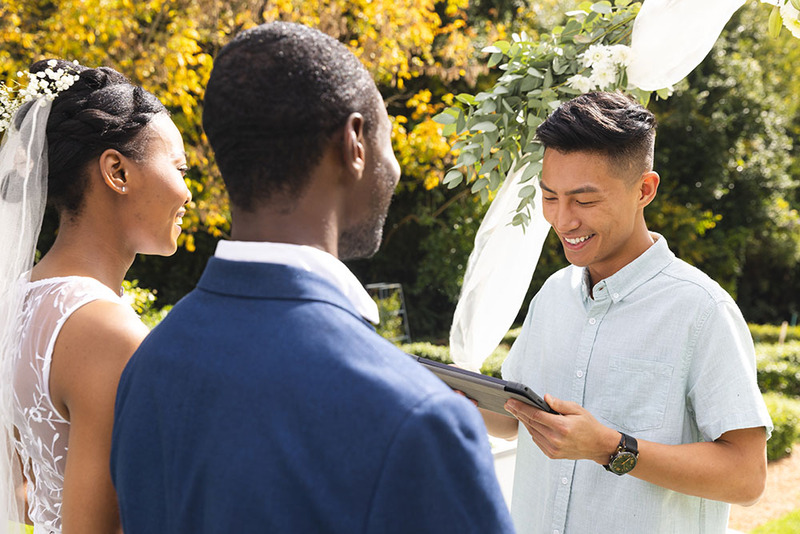 An ordained minister officiates a wedding for a bride and groom outdoors in the sunshine on a summer day. The officiant is looking down at the wedding script as he smiles and reads the ceremony