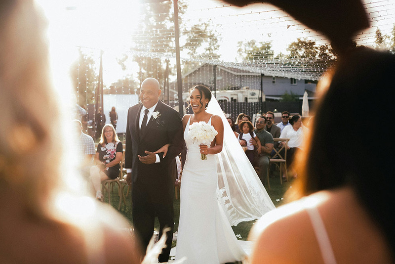 A bride and her father walk down the aisle at the start of the wedding ceremony while friends and family members cheer