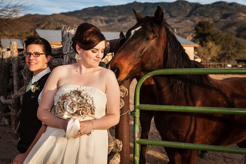 Lesbian couple on their wedding day in a beautiful outdoor ranch setting, they pose next to a horse in their wedding outfits, looking lovely and happy