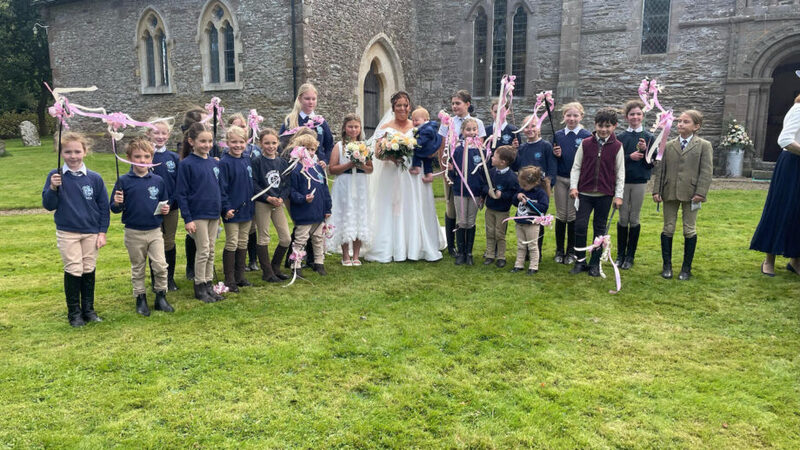 Photo via Horse and Hound, shows children holding up riding crops decorated with pink ribbons, standing next to a bride on her wedding day. 