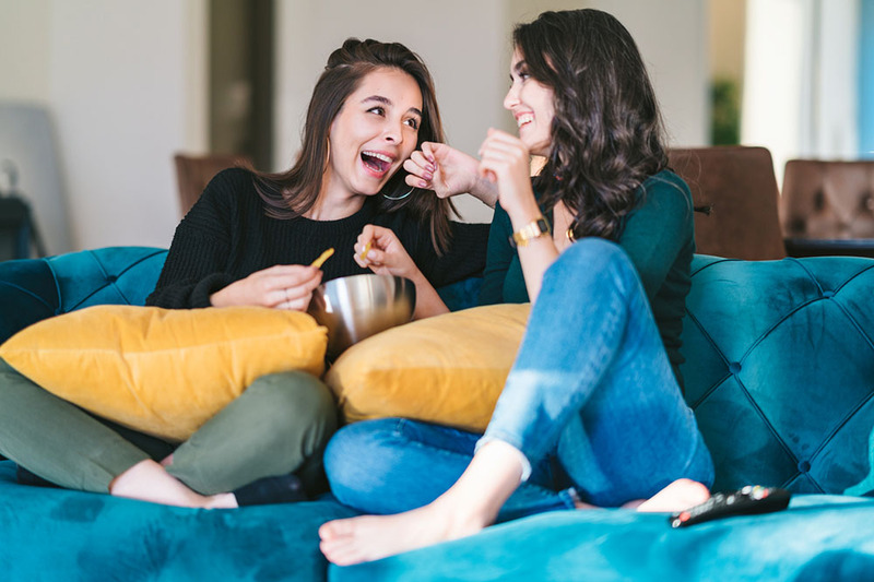 Two women sit on a couch eating snacks and laughing together, to symbolize modern lavender marriage.