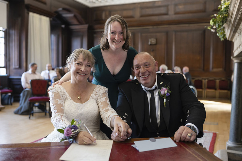 A wedding officiant poses with the bride and groom after their ceremony while they sign the marriage license