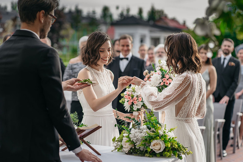 Two brides exchange wedding rings during an outdoor wedding ceremony with a wedding officiant and friends and family members