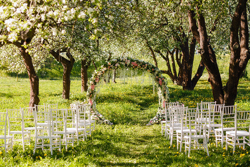 A spring wedding scene outdoors with white blossoms on the trees and chairs set up in front of a wedding arch before the ceremony