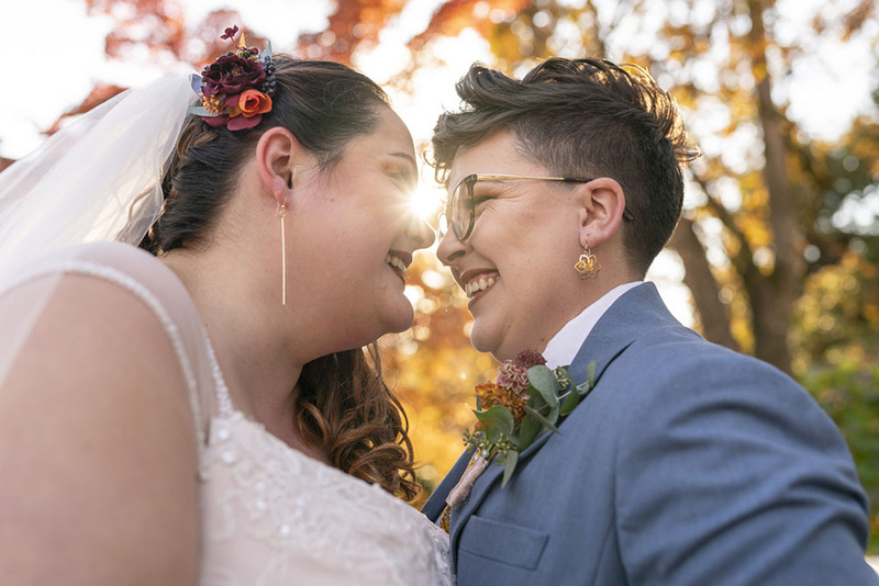 Close up of brides smiling happily on the wedding day in their wedding attire, in the background are colorful fall leaves on trees and some sun shining through