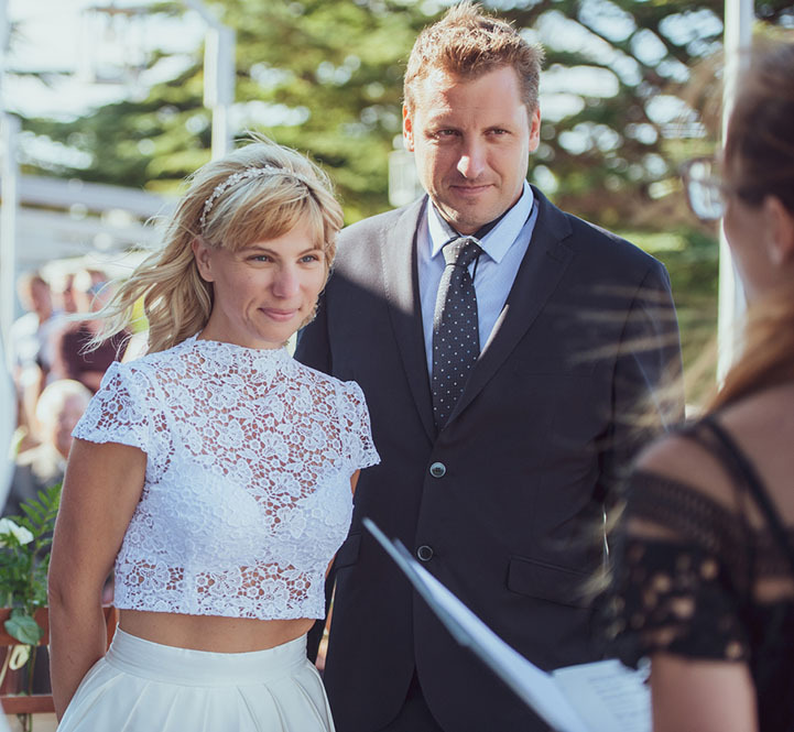 Bride and groom standing in front of a wedding officiant, listening and smiling as the officiant performs the ceremony. In the background, they are outdoors on a sunny day. There are green trees, and chairs set up for guests and family