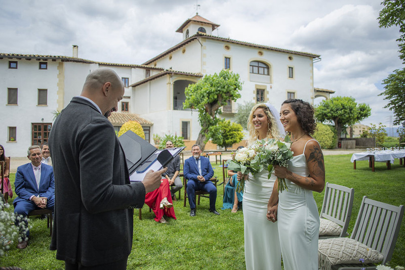 A wedding officiant reads from a wedding ceremony script during an outdoor ceremony, two brides stand in front of him holding hands and smiling. They are watched by a group of friends and family in nice colorful clothes