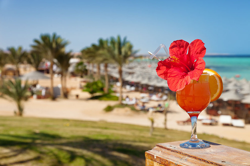 A hibiscus cocktail sits on a table outdoors in the sun with the beach in the background