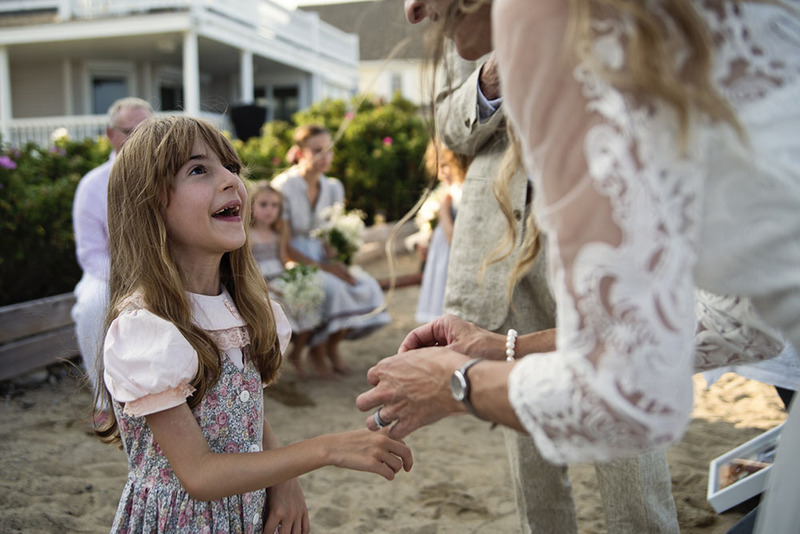 A young girl looks up at her mom with excitement, the mom is wearing a white wedding dress for her elopement. In the background are older family members, the dad / groom, and other children