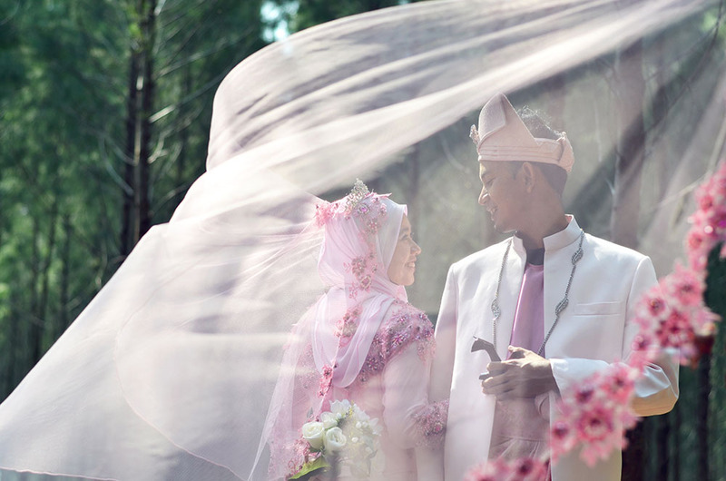 Bride and groom pose for wedding photos outdoors. The woman is wearing traditional Muslim bridal wear in soft pinks, and the veil falls in front of her and the groom as they smile at each other lovingly