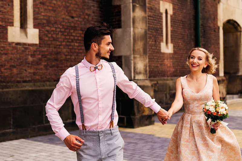 Groom holds hands with the bride as they laugh and run down the street happily. The groom wears a pink dress shirt and the bride wears a peachy pink patterned dress. 