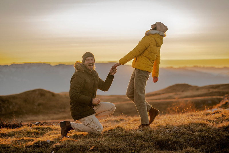 Outdoor photo shows man on one knee, proposing marriage to a woman. She is laughing and happy, she said yes!