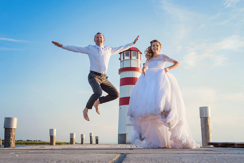 Bride and groom jump with joy in front of a lighthouse after their beach wedding