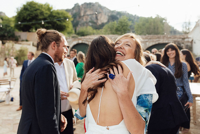 Family member wedding officiant hugs the bride after the wedding ceremony. They look overjoyed! Friends and relatives walk around in the background outdoors on a sunny day
