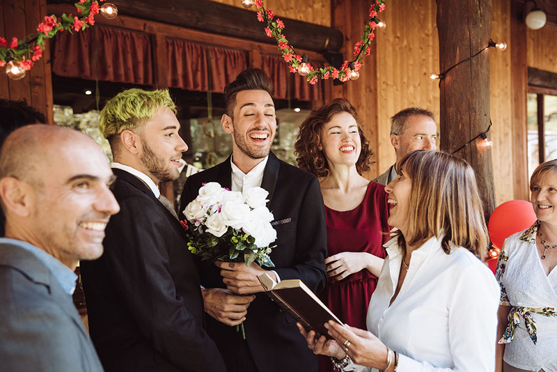 Two grooms stand together on the wedding day with their officiant, friends, and family. Everyone is happy!