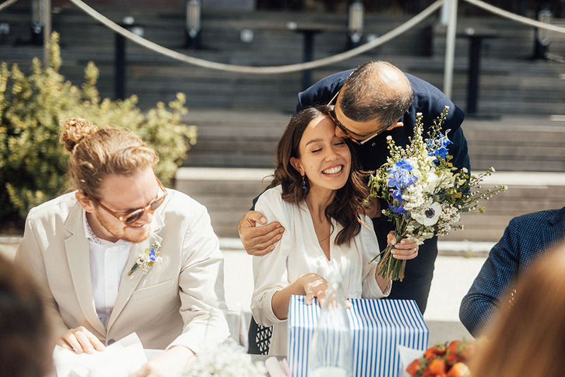 Groom hugs a seated bride as she opens wedding gifts on a sunny day during the wedding reception