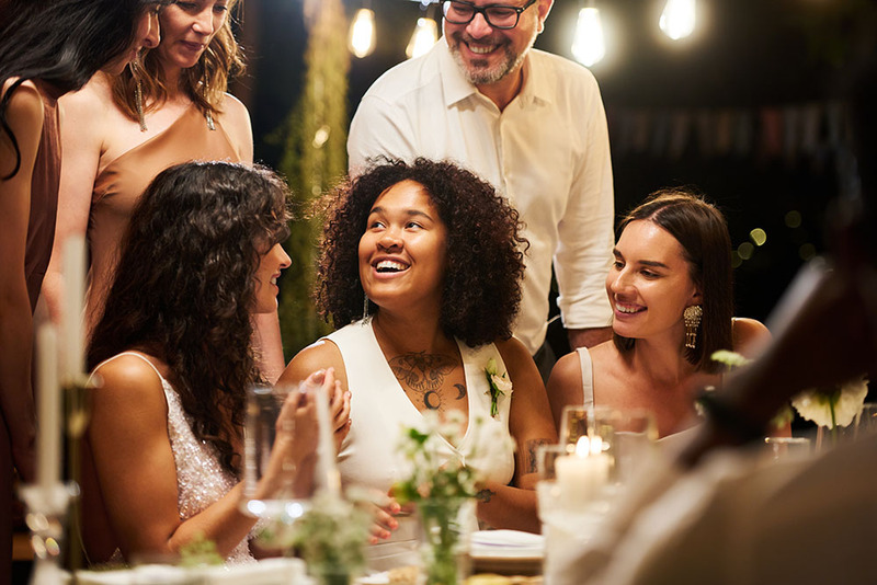 Group of friends stand next to the two brides, seated at the wedding reception dinner. 
