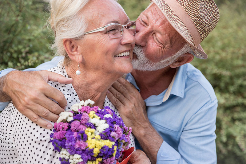 An older couple on their wedding day, embracing and smiling, the groom kisses the bride on the cheek