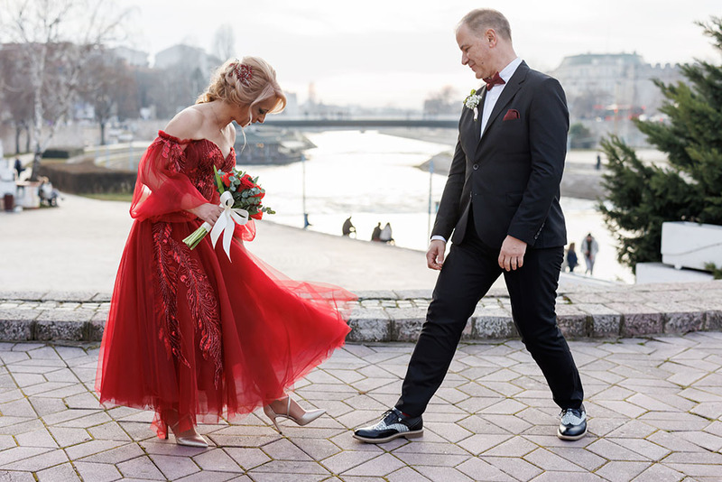 A bride in a red wedding dresses stands with a groom in a dark suit, they are showing off their shoes