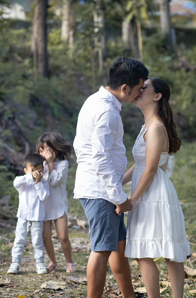 Una joven pareja casada se besa en la renovación de sus votos mientras sus hijos miran al fondo, sonriendo y actuando tímidos, cubriéndose los ojos.