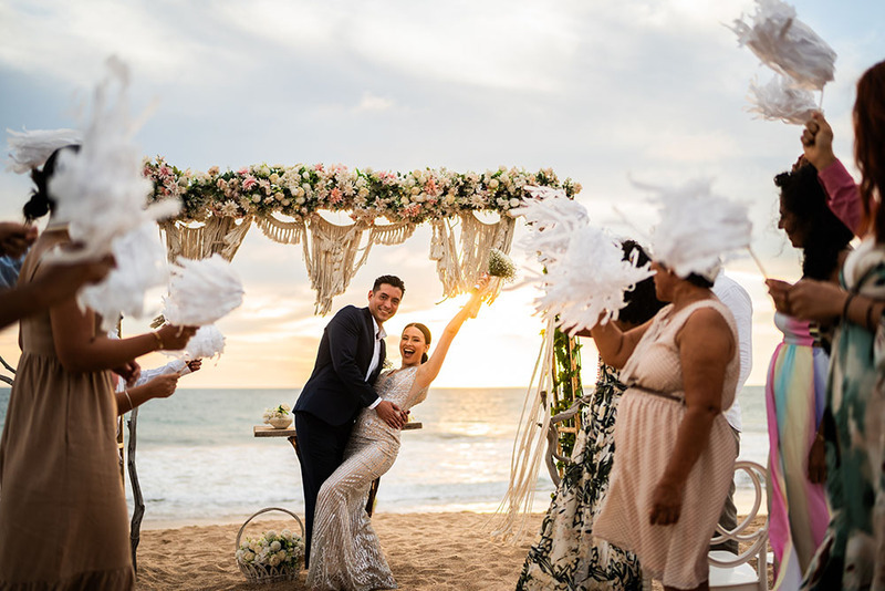 Los novios posan al final de su ceremonia nupcial mientras amigos y familiares los animan. En la playa al atardecer.