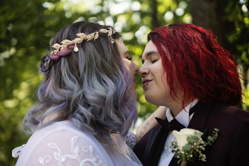 Two brides embrace happily on the wedding day; one has blue hair with a decorative head wreath, the other has red hair and is wearing a bowtie