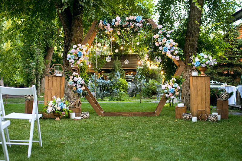 A beautiful wedding arch with flowers is set up in a backyard, with chairs and decor. There are green leafy trees, bushes, and a house in the distance.