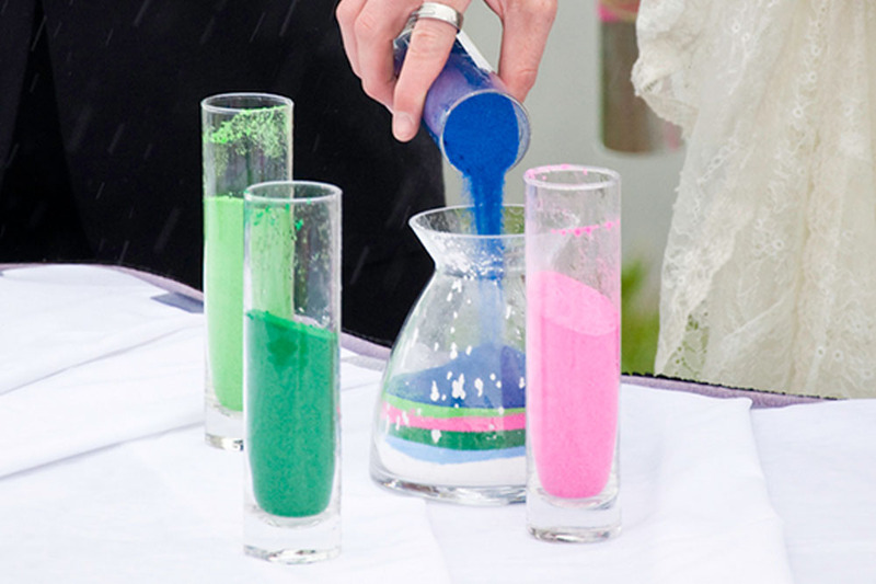 Close up image of a unity sand ceremony at wedding, as groom pours blue sand into a vase. Table holds more containers, with different colored sand (greens and pink too)