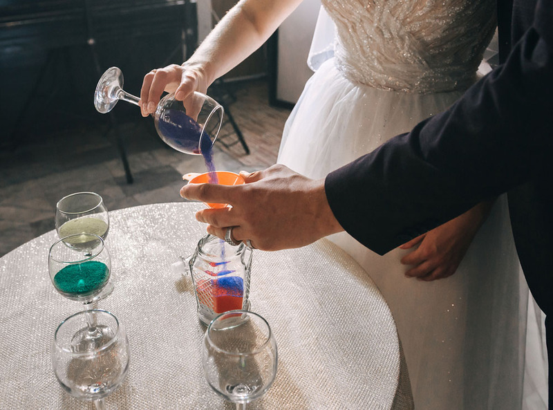 Close up of bride pouring colored sand during a wedding unity sand ceremony, wine glasses hold different colors of sand on the table, and the groom holds a funnel while she pours to help the sand go into the vase
