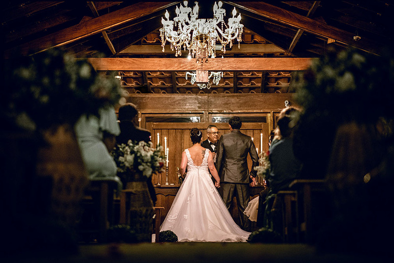 A bride and groom are married in a church by a wedding officiant, there are lovely dark wood accents and exposed beams along the ceiling, and warm lighting for a cozy peaceful vibe