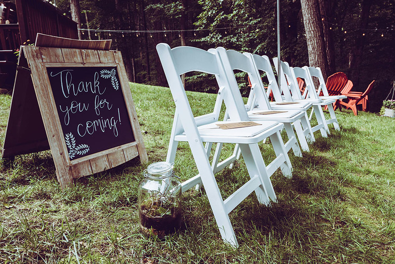 A backyard wedding celebration is set up, with white folding chairs arranged in the grass and a wedding welcome sign with 'thank you for coming!' written in cursive in chalk