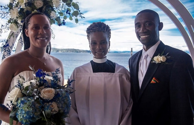 Rev. T, Minister Tiji Murphy, poses with newlyweds after performing their wedding, they all smile happily at the camera against a decorative wedding arch