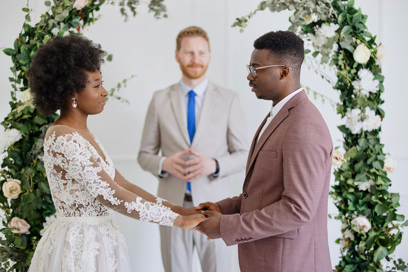 bride and groom hold hands in front of a decorative wedding arch, exchanging marriage vows, while the wedding officiant smiles in the background happily