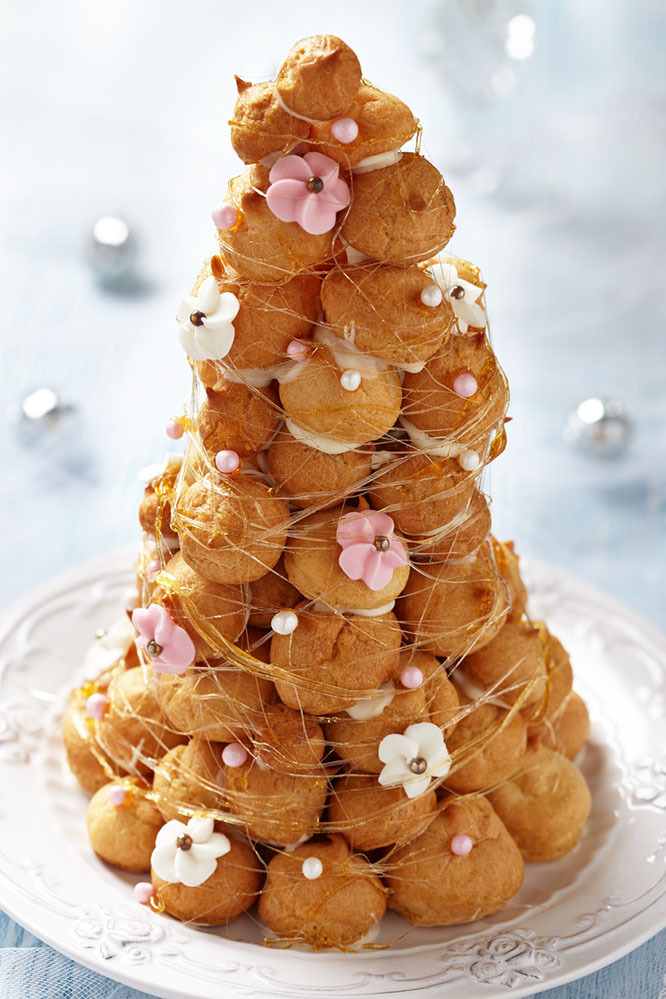 A beautiful example of a croquembouche on a white table, with puff pastries covered in thin threads of caramel and flowers made of pink and white icing